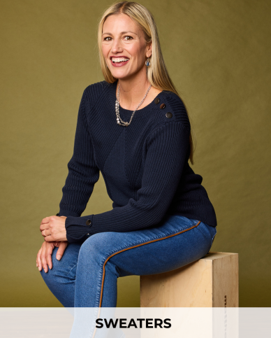 A smiling blonde woman sits on a wooden block wearing a dark navy ribbed sweater and blue jeans against an olive green studio backdrop. A white banner at the bottom reads 'SWEATERS'.