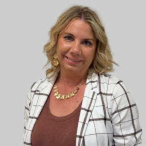 A woman with blonde hair wearing a brown top, a white blazer with a brown grid pattern, and gold jewelry, standing against a plain off-white wall.